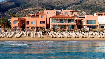 Un hotel costero con varias construcciones, una playa con tumbonas y un telón de fondo montañoso.
