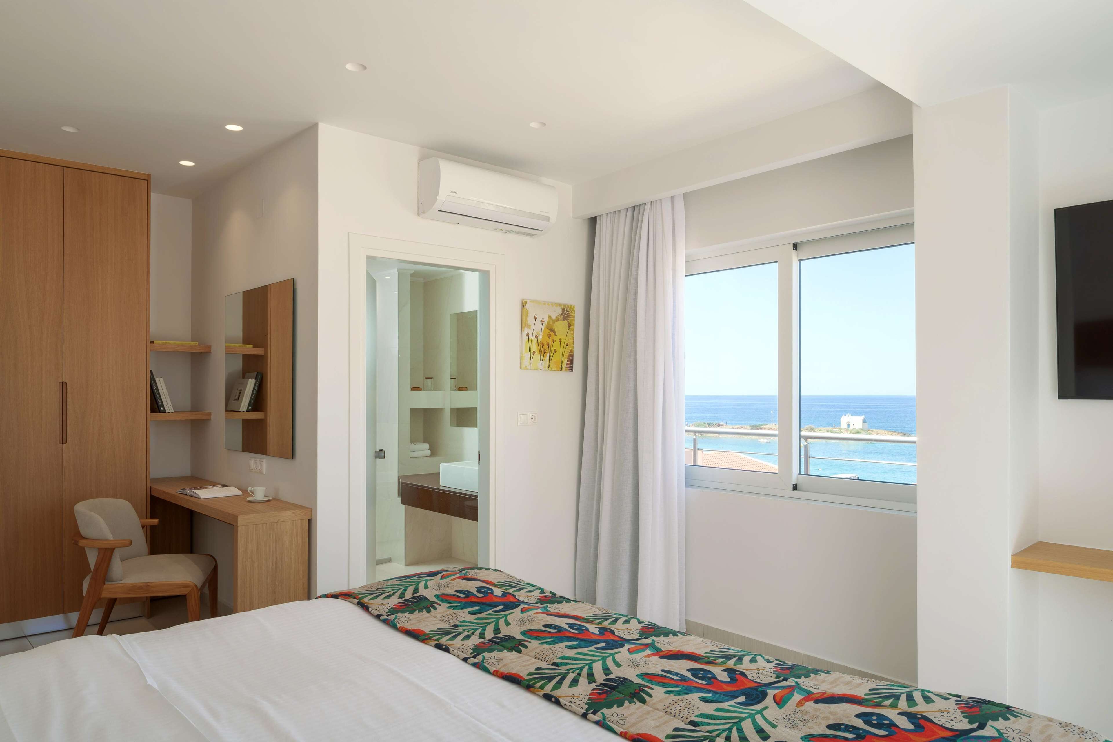 A bedroom with a bed, a desk, a chair, a wardrobe, a mirror, and a window with a view of the sea.
