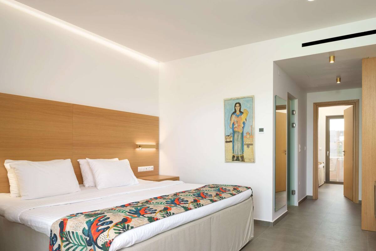 A modern hotel room with a wooden headboard, a bed with white linens, a colorful bedspread, and a painting on the wall.