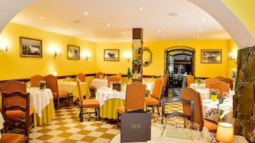 Un restaurant amb parets grogues, un terra de taules de viqueta i cadires de fusta. Taules cobertes amb teles de taula blanques i copes de vi.