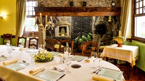 A dining area with a stone fireplace, wooden beams, and a table set for four with wine glasses and plates. - Le Vieux Logis (Lestelle-Betharram)