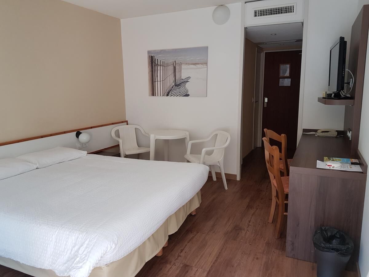 A hotel room with a bed, a desk, a chair, a small table, and a picture on the wall.