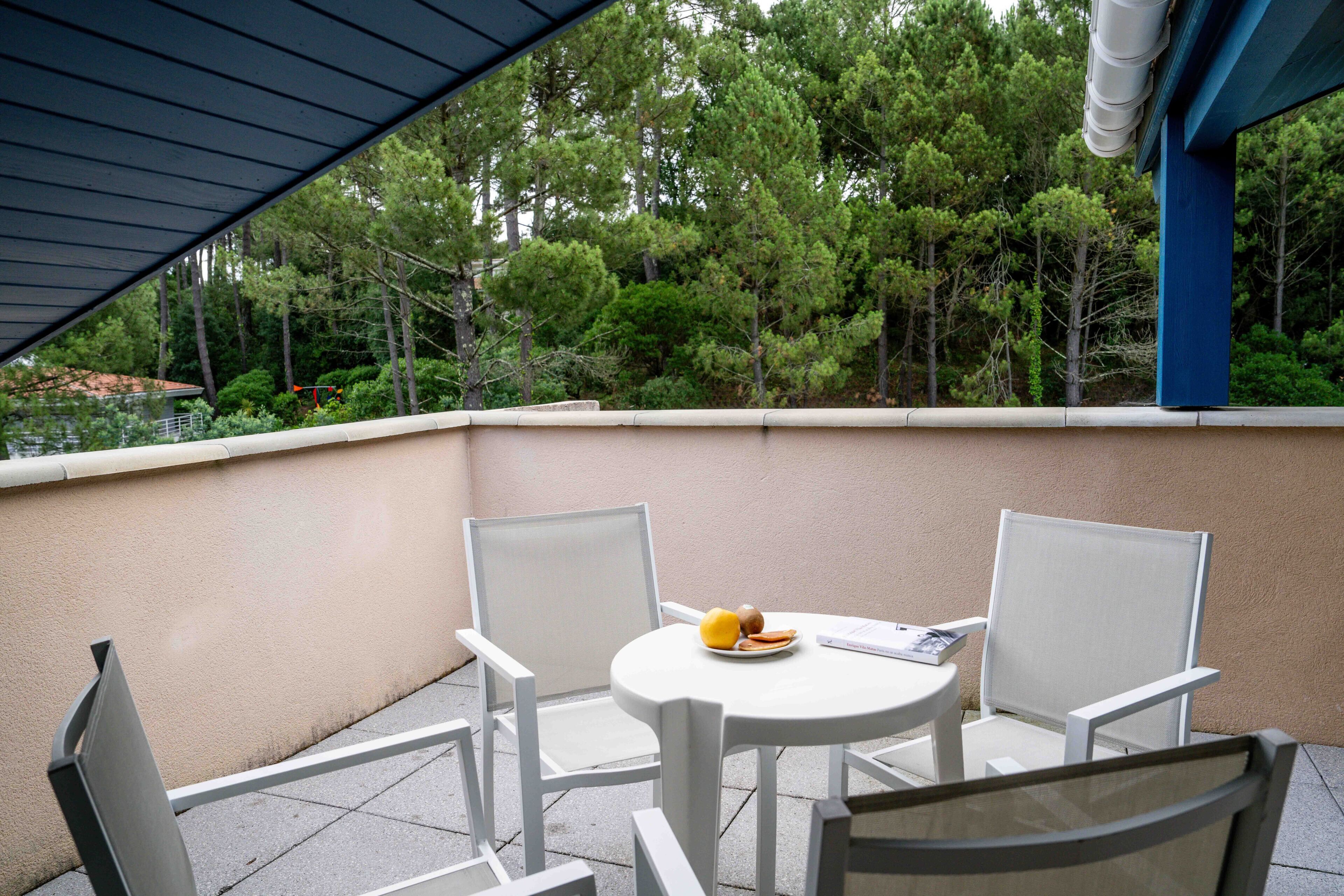 Een balkon met twee witte stoelen en een kleine tafel, een blauw zonnescherm en een uitzicht op bomen en een gebouw op de achtergrond.