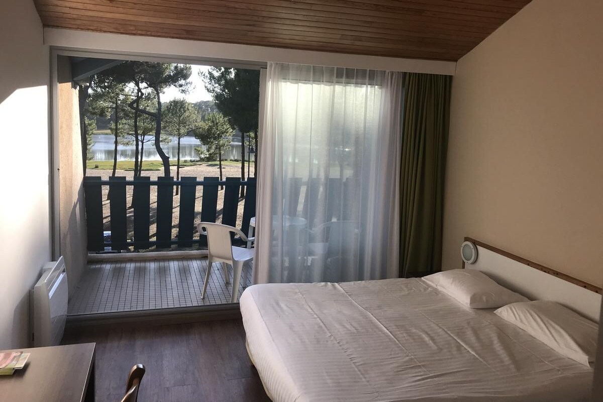 Quarto de hotel com cama, escrivaninha, cadeira, janela com cortinas e vista para o exterior.