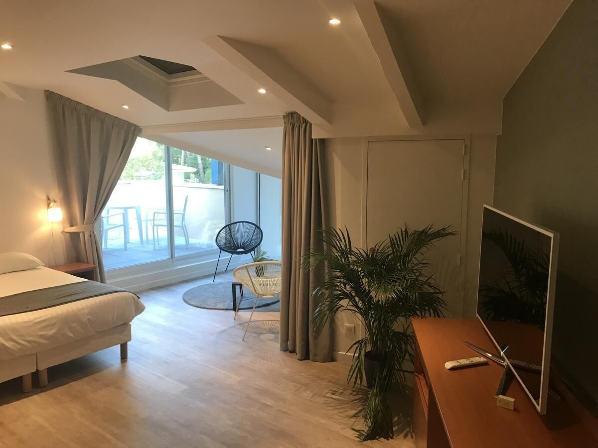 A modern hotel room with a bed, a desk with a TV, a chair, a potted plant, and a balcony with outdoor seating.