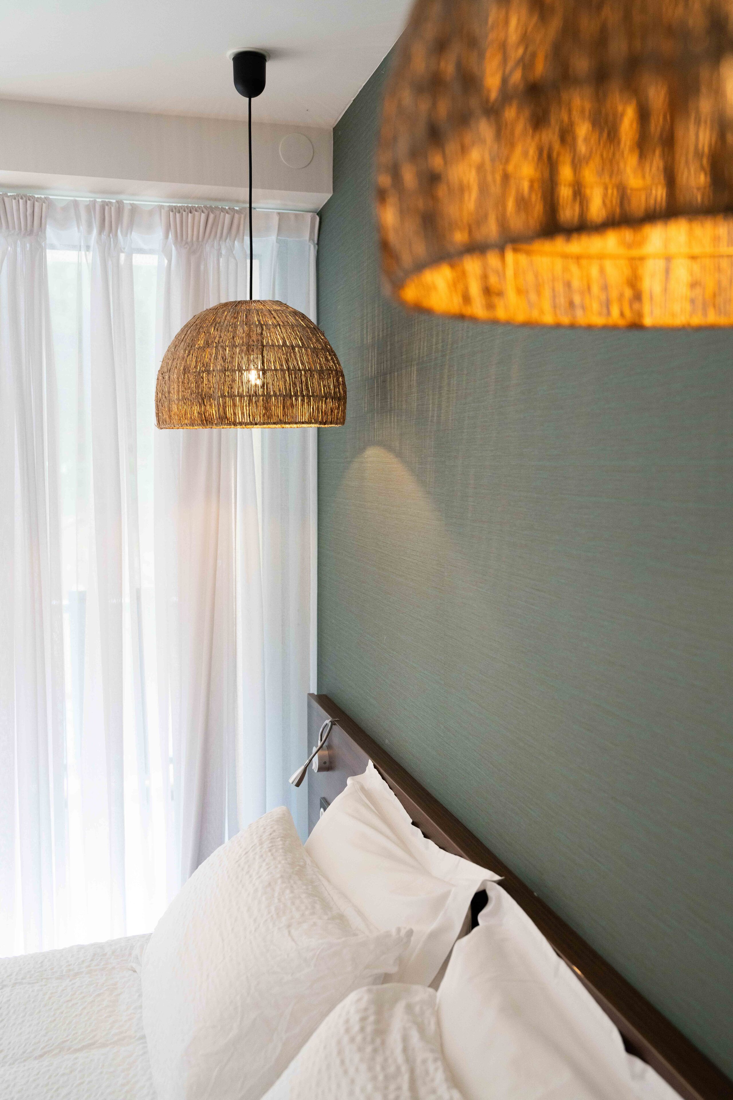 A hotel room with a bed, a hanging woven light fixture, and sheer curtains.