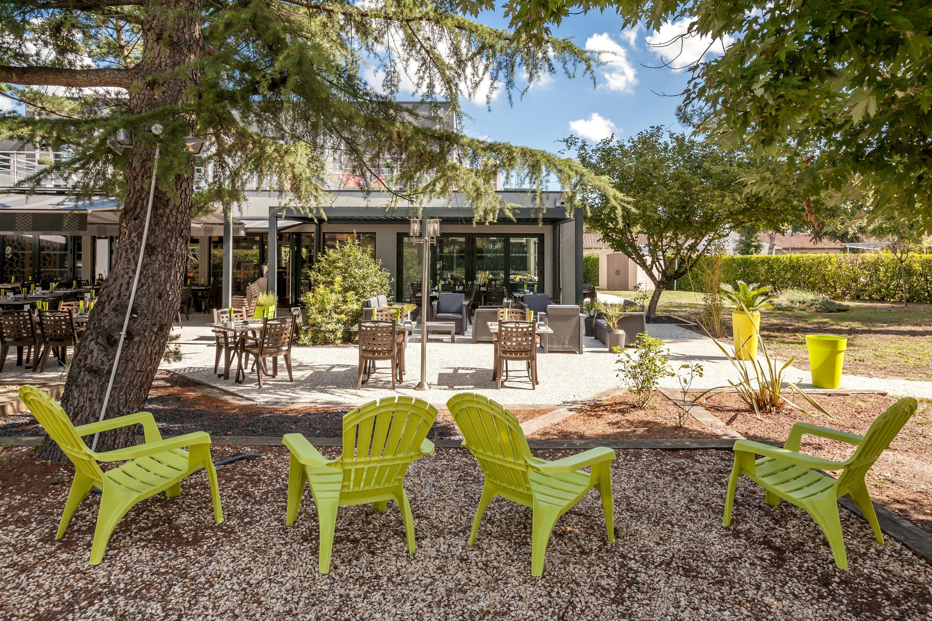 An outdoor seating area with yellow chairs and tables, surrounded by trees and a building with large windows.