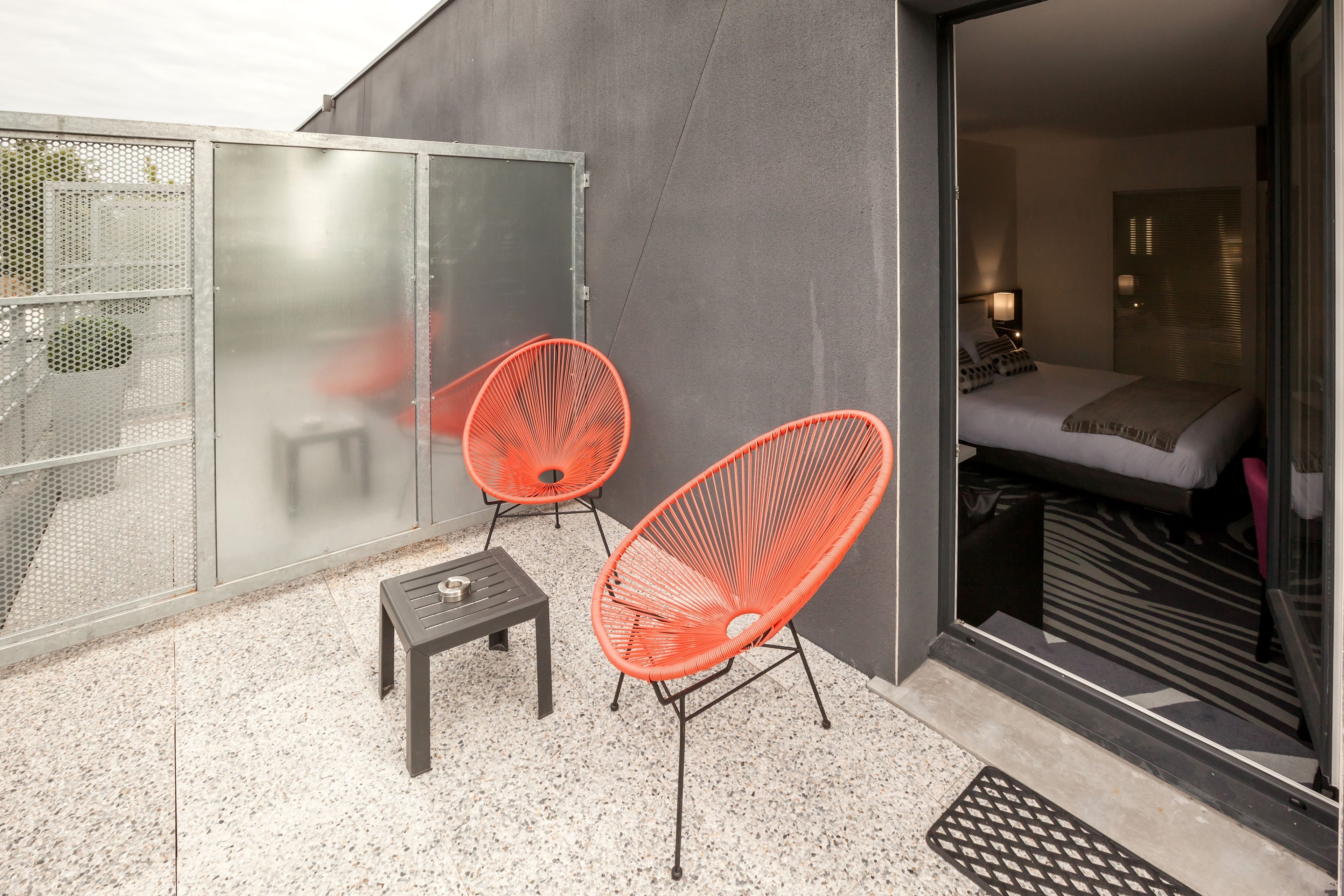 A modern hotel room with a bed, a small table, two orange chairs, and a glass door leading to the outside.