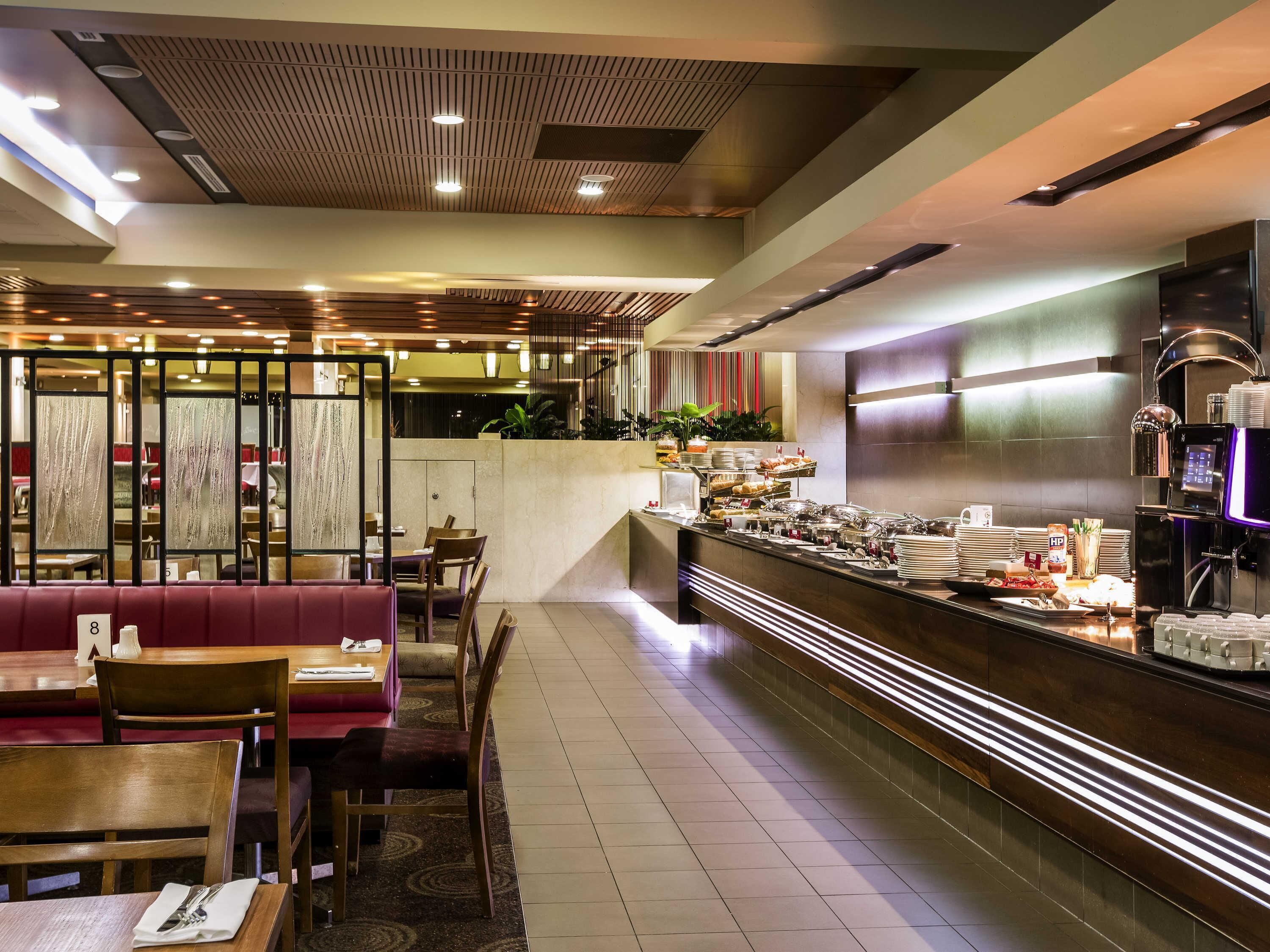 Comedor de hotel con barra libre, mesas y sillas, y zona de cocina.