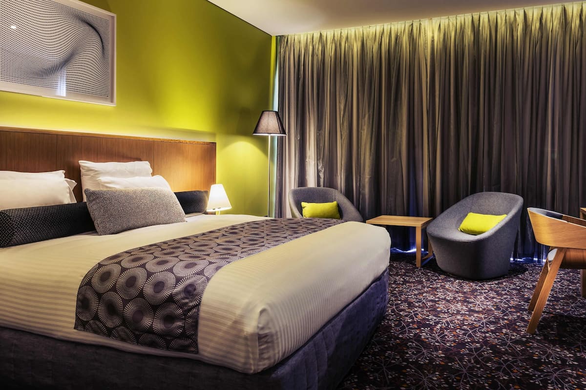 A hotel room with a large bed, a headboard, a bedside table with a lamp, two chairs, a small table, and a large window with curtains.