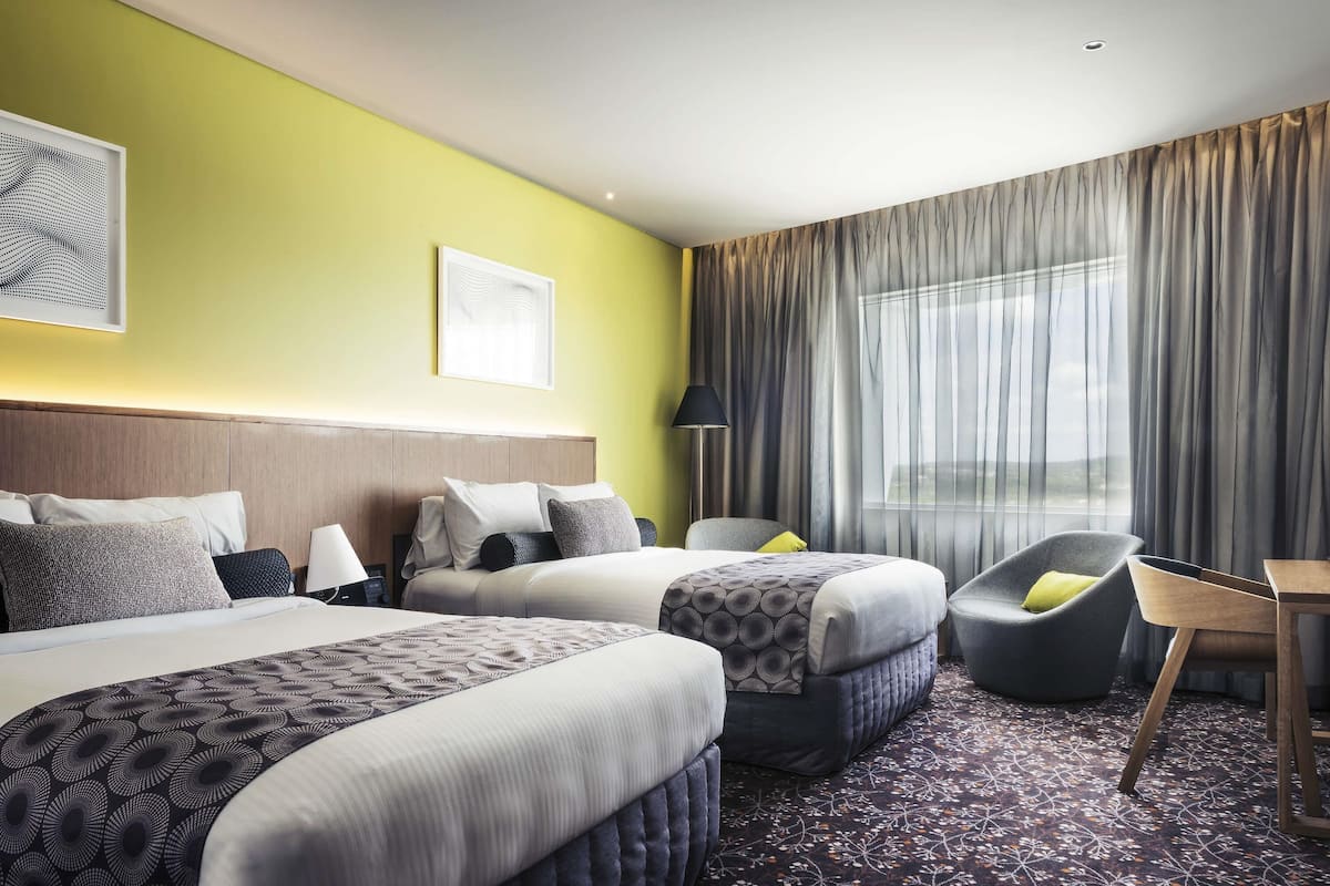 A hotel room with two beds, a chair, a table, and a large window with curtains.