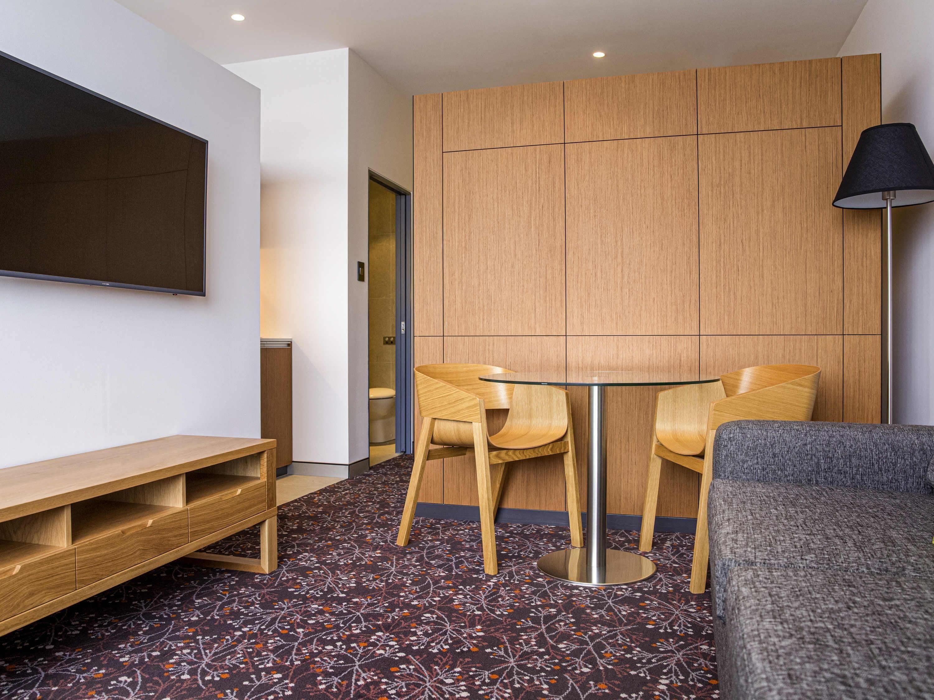 Une chambre d’hôtel avec une armoire en bois, une table en verre avec des chaises, un canapé, un téléviseur fixé au mur et une moquette à motifs.