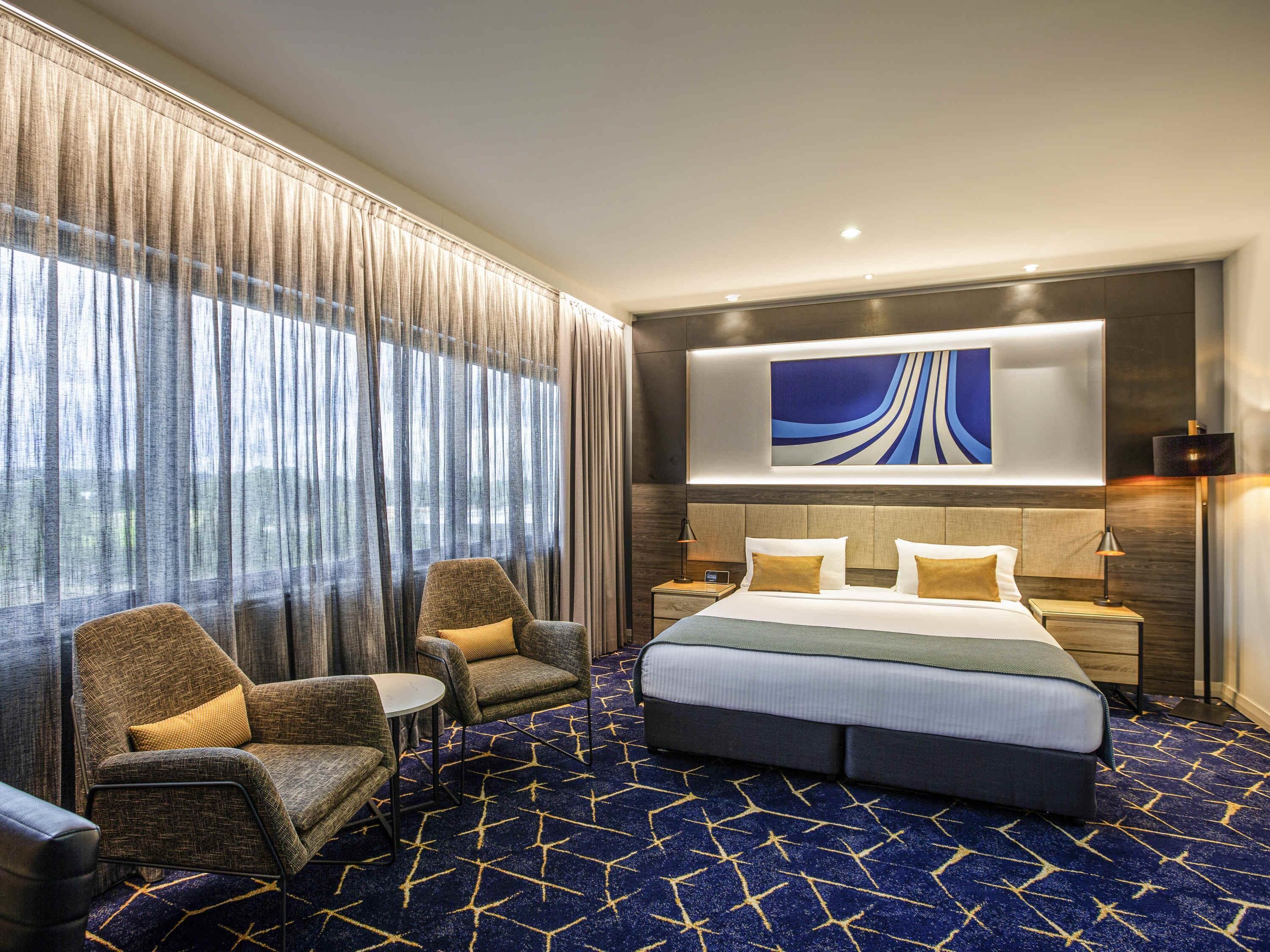 A hotel room with a large bed, two armchairs, a small table, a nightstand, a wall-mounted artwork, and a view through sheer curtains.