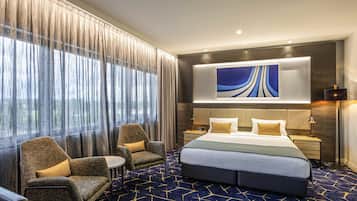 A hotel room with a large bed, two armchairs, a small table, a nightstand, a wall-mounted artwork, and a view through sheer curtains.