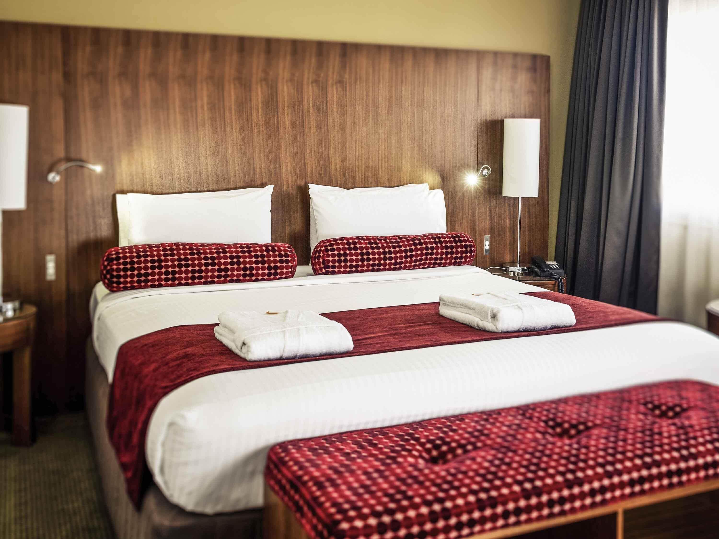 A hotel room with a wooden headboard, two white pillows, a red and black patterned pillow, a red throw blanket, and a white towel on the bed.