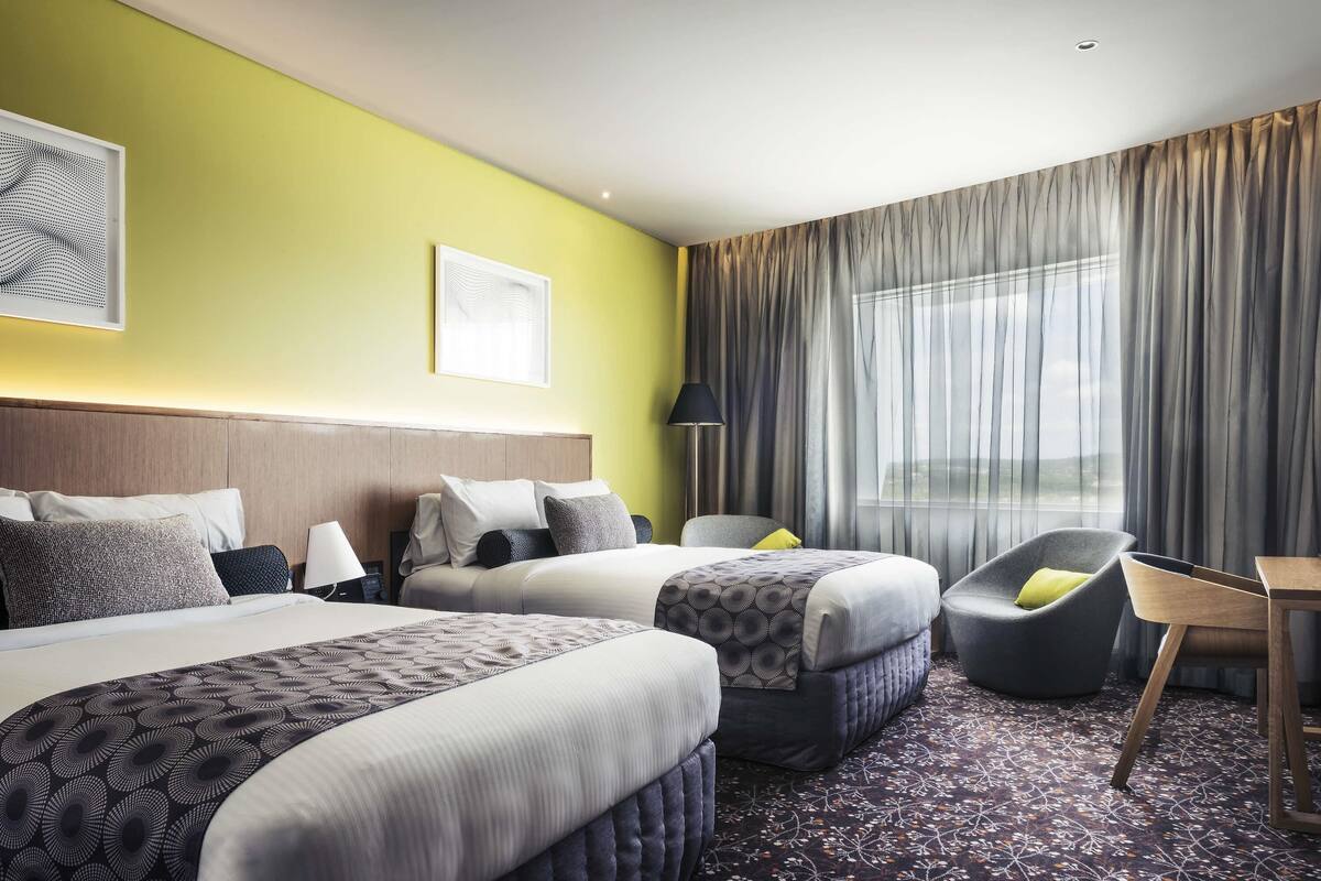 A hotel room with two beds, a chair, a table, and a large window with curtains.