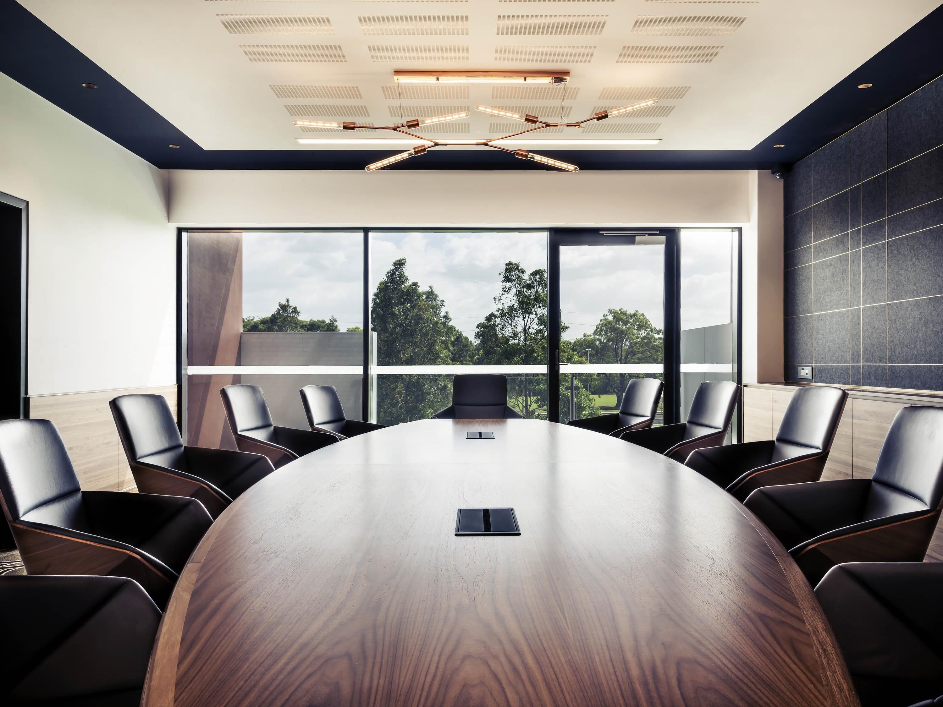 Un salón de conferencias con una gran mesa de madera, sillas de cuero negro y ventanas amplias con vistas a árboles.