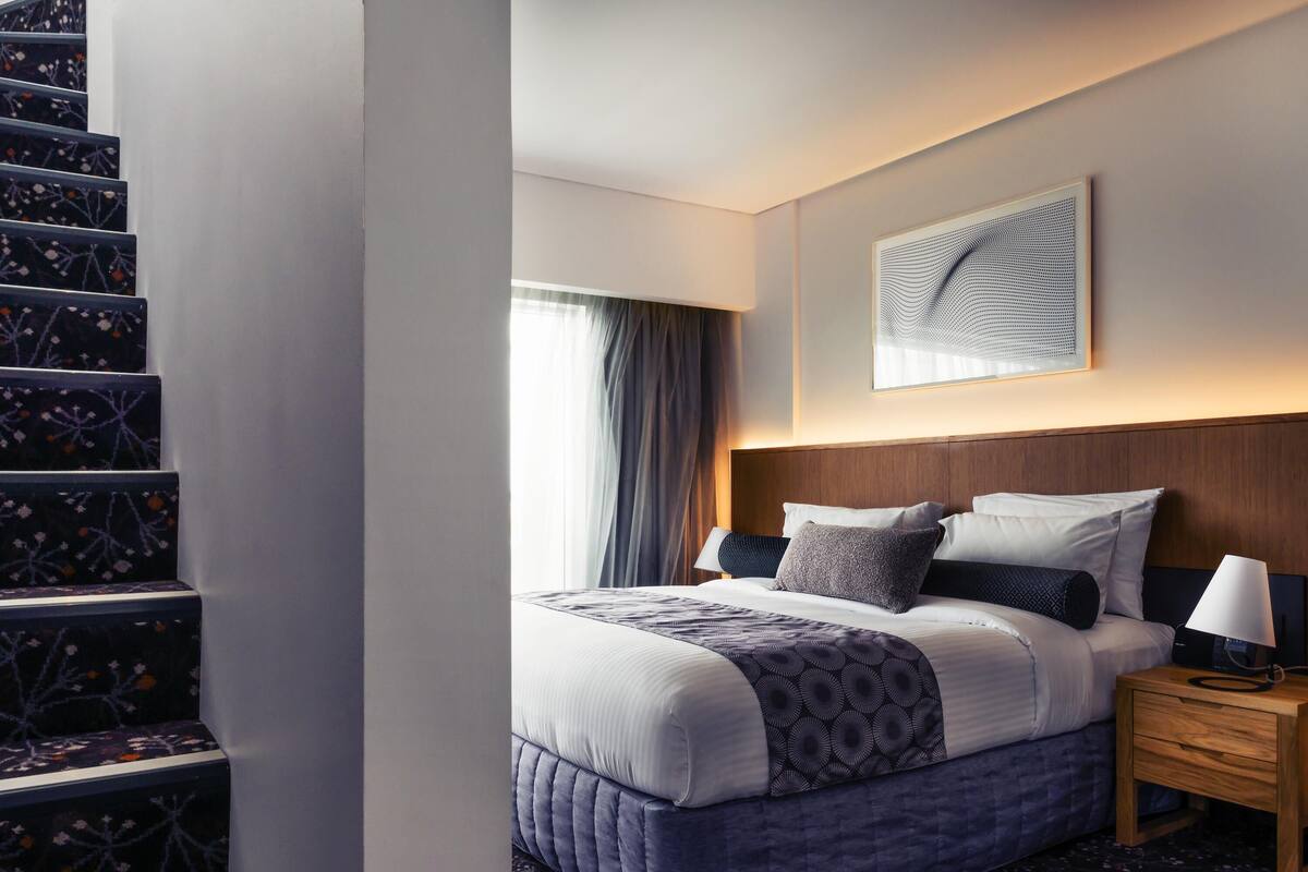 A hotel room with a bed, a nightstand, a wall-mounted artwork, and a staircase with floral-patterned steps.
