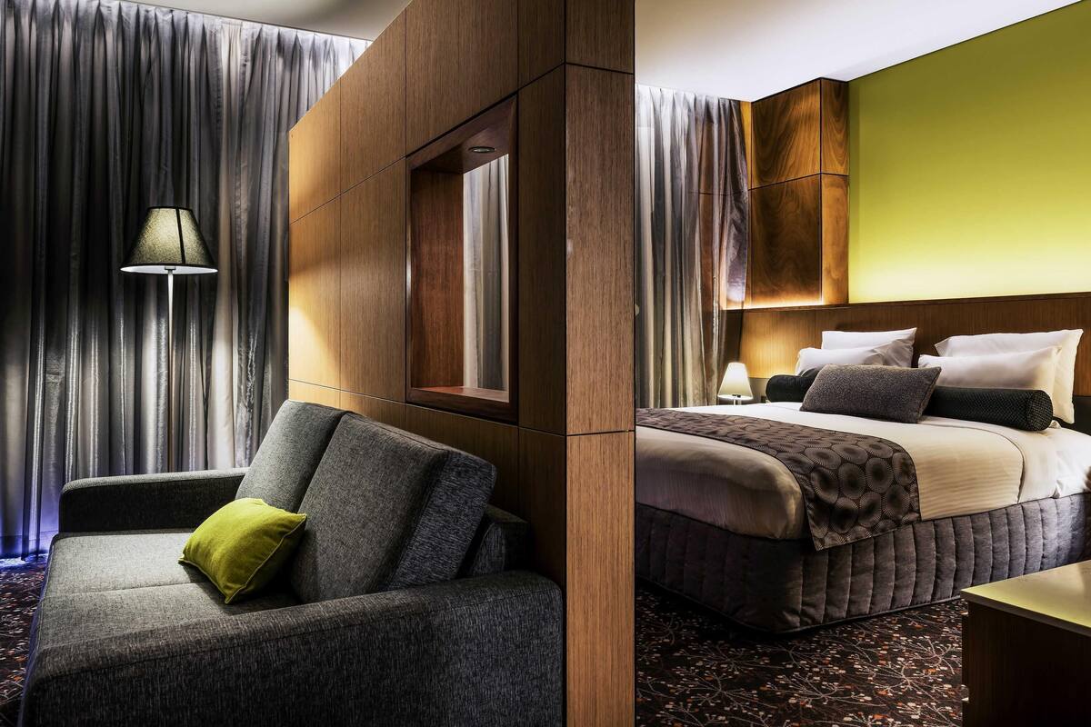 A hotel room with a sofa, a bed with pillows, a lamp, and a large window with curtains.