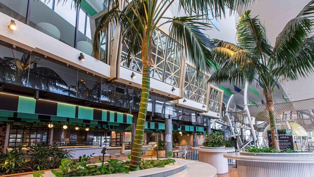 A modern indoor atrium with a central circular seating area, multiple levels with glass railings, and lush greenery including palm trees. - Mercure Sydney Liverpool (Prestons)