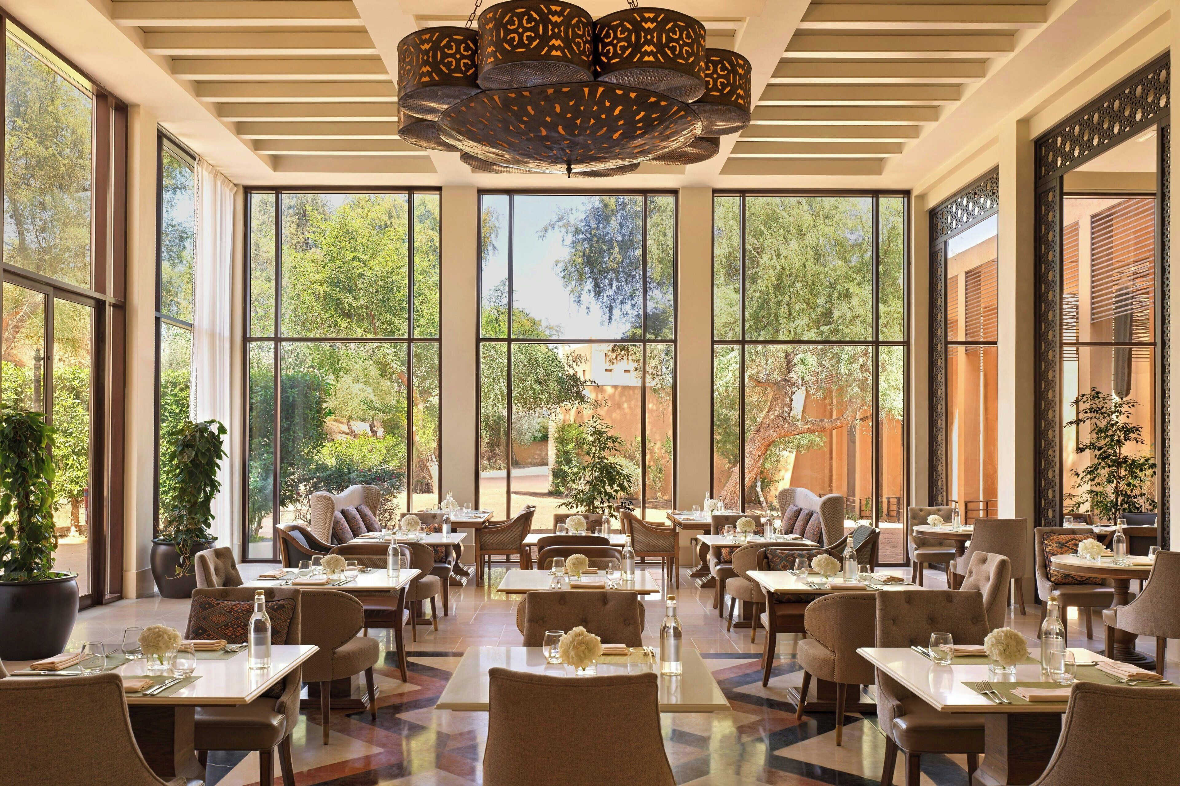 A spacious dining area with large windows, multiple tables, and chairs, featuring a modern chandelier and potted plants.