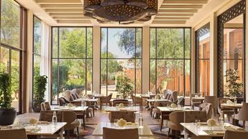 A spacious dining area with large windows, multiple tables, and chairs, featuring a modern chandelier and potted plants.