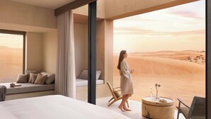 A modern hotel room with a large bed, a seating area with cushions, a round wooden table with a vase and bottle, and a view of desert dunes through large windows. - The Ritz-Carlton Ras Al Khaimah, Al Wadi Desert (Ras Al Khaimah)