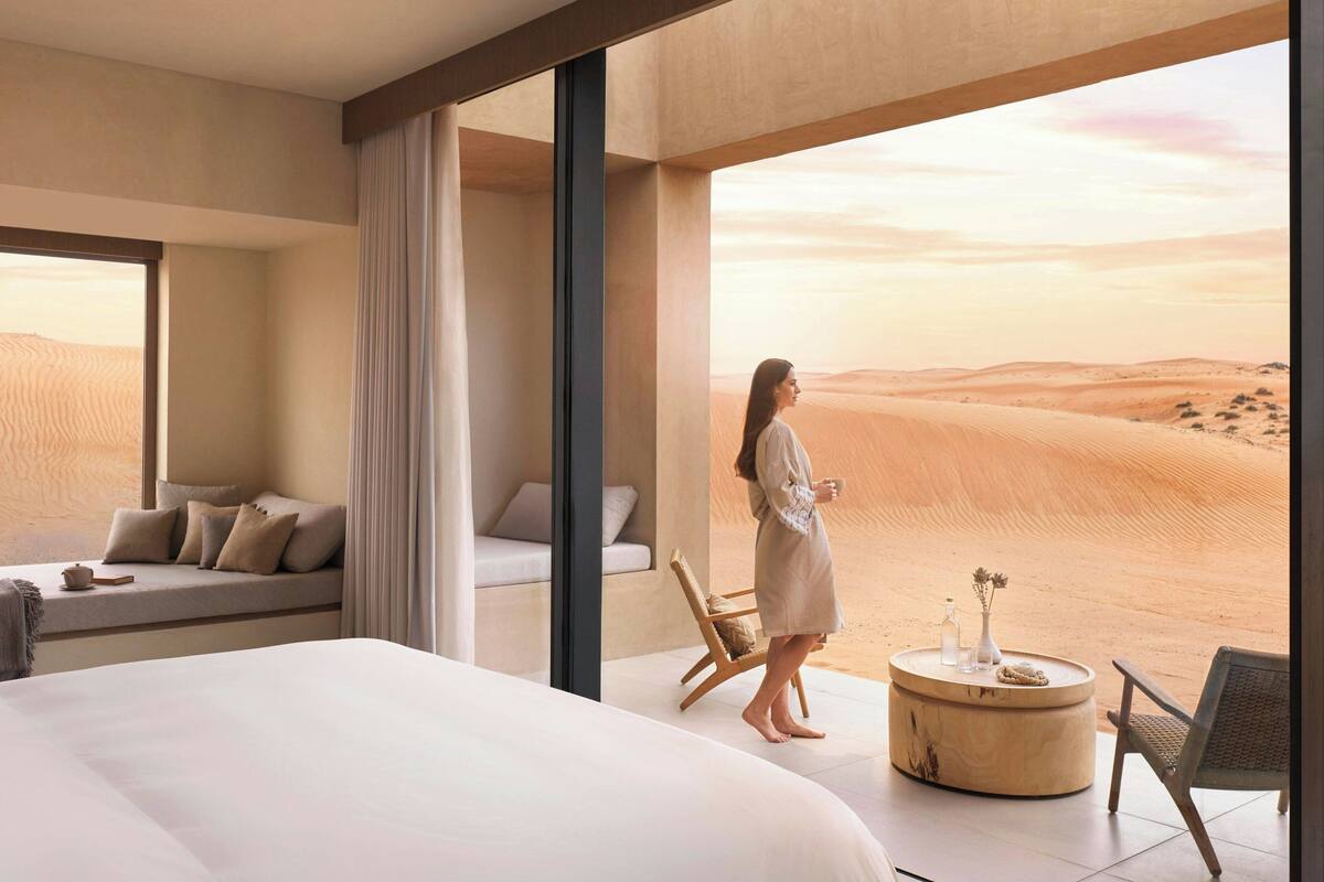 Ett modernt hotellrum med en stor säng, en sittgrupp med kuddar, ett runt träbord med en vas och en flaska, och utsikt över ökenlandskapet genom stora fönster.