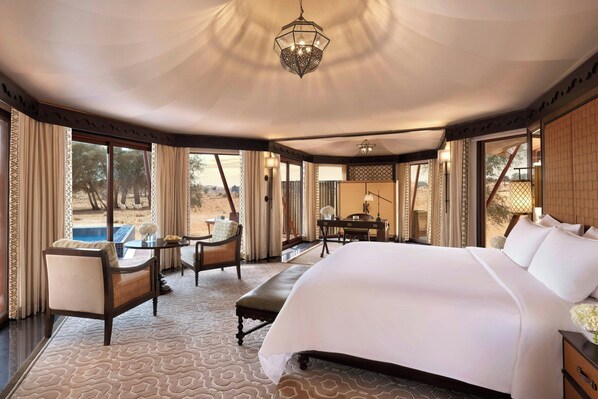 A spacious bedroom with a large bed, a desk, and a chair. There is a view of the outdoors through large windows.