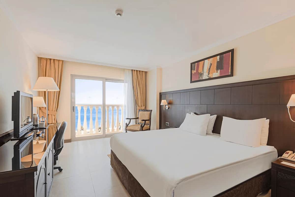 Una habitación de hotel con una cama grande, un escritorio con una computadora, una silla, una mesita de noche con un teléfono y un balcón con vistas.