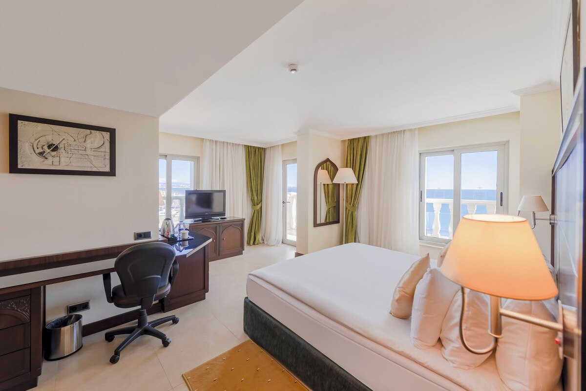 A hotel room with a bed, desk, chair, TV, and windows.