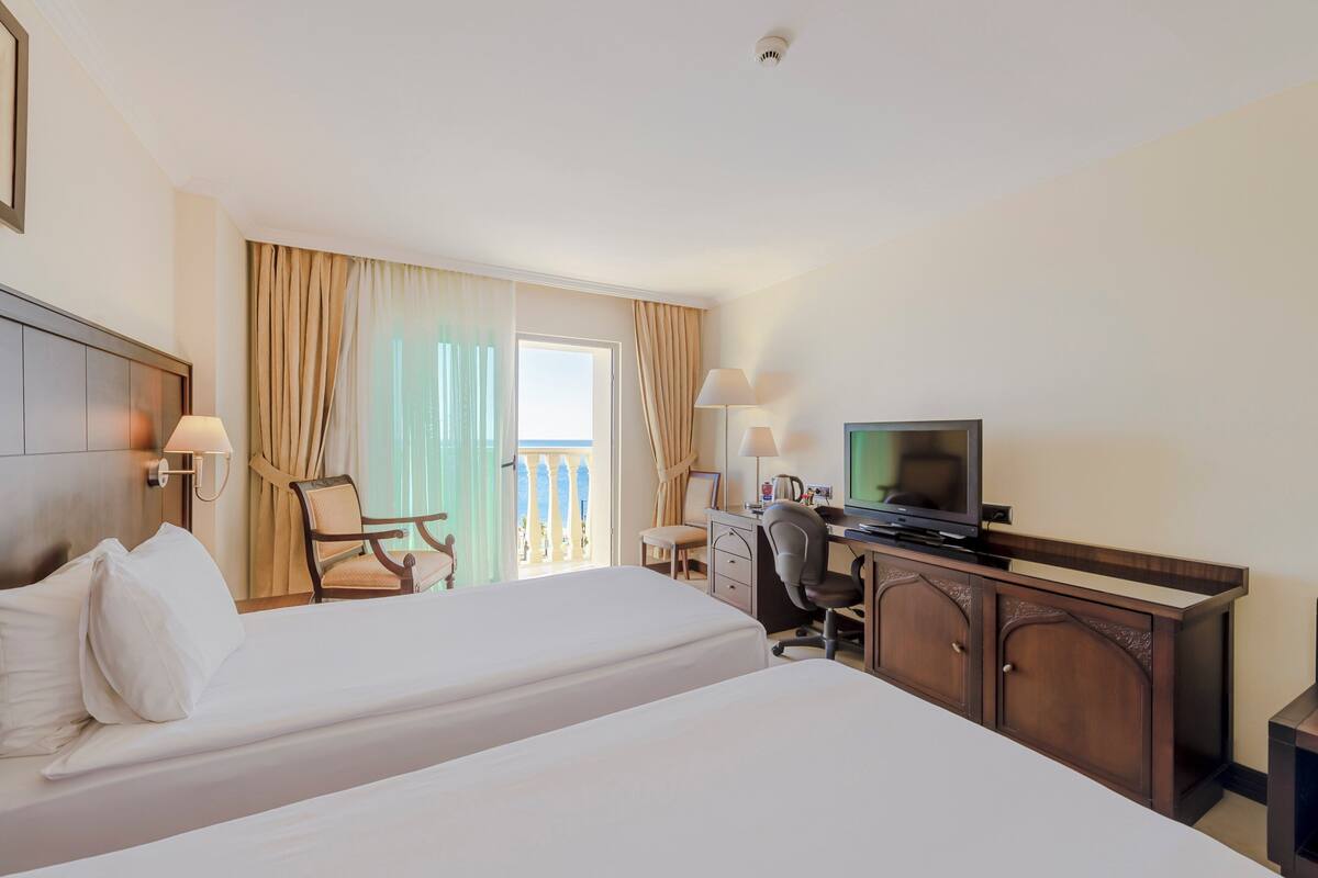 Habitación de hotel con dos camas, un escritorio con televisor, una silla y un balcón con cortinas.