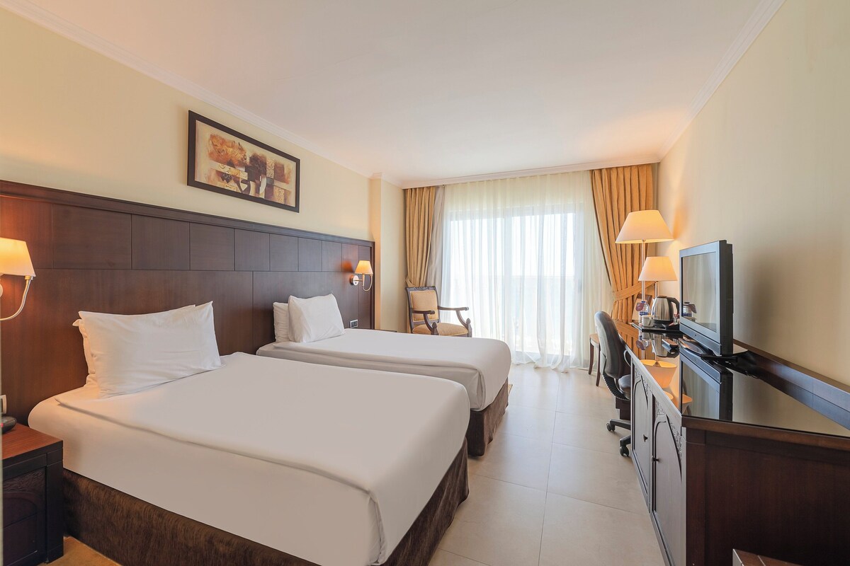 A hotel room with two beds, a desk with a TV, a chair, and a window with curtains.