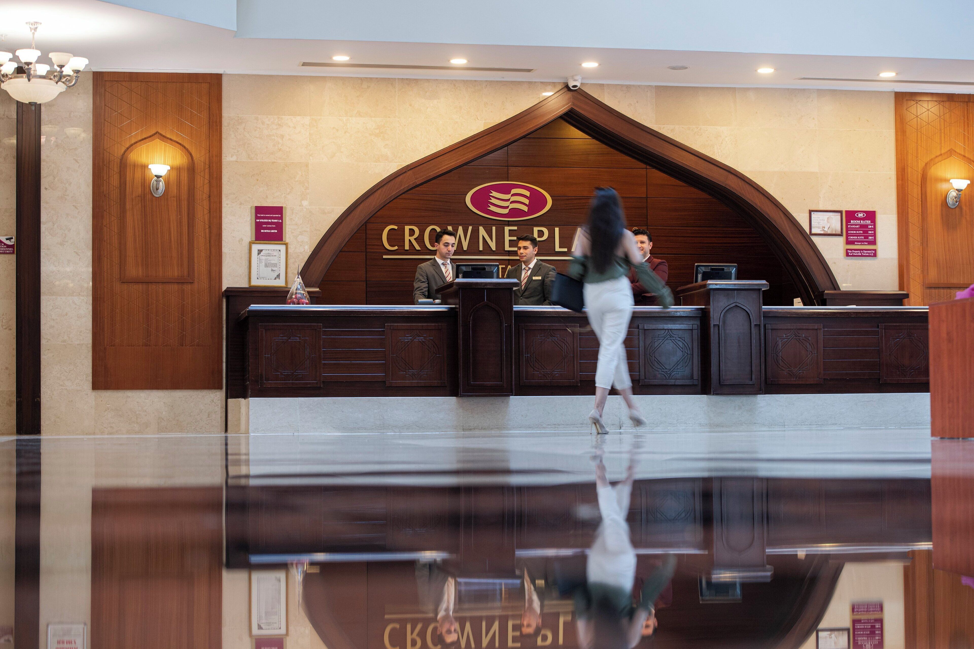A hotel reception area with a wooden reception desk, a large logo above the desk, and a glass barrier reflecting the interior.