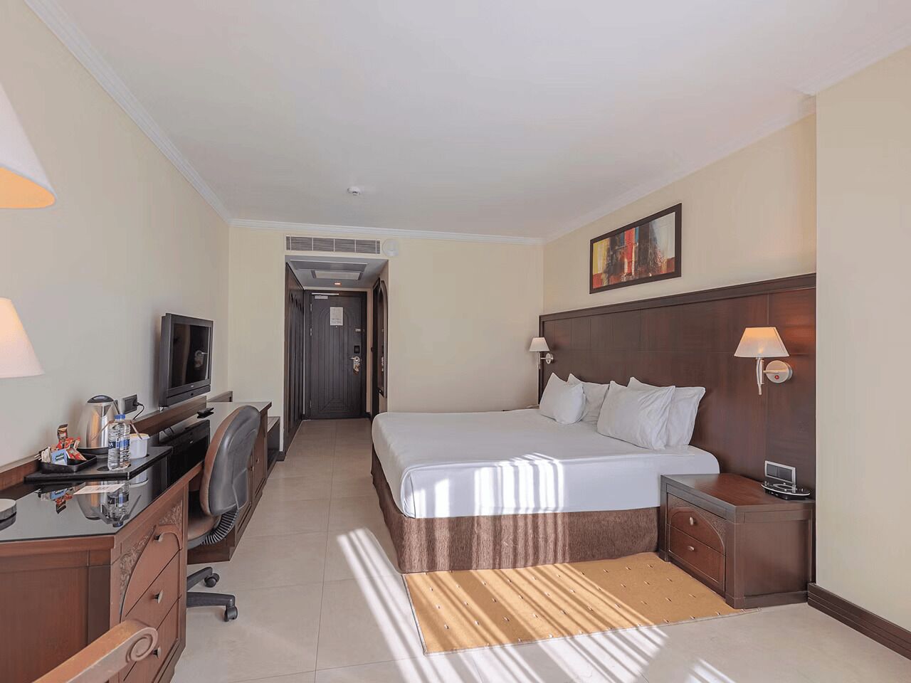 A hotel room with a bed, bedside tables, a desk with a computer, a TV, and a lamp.