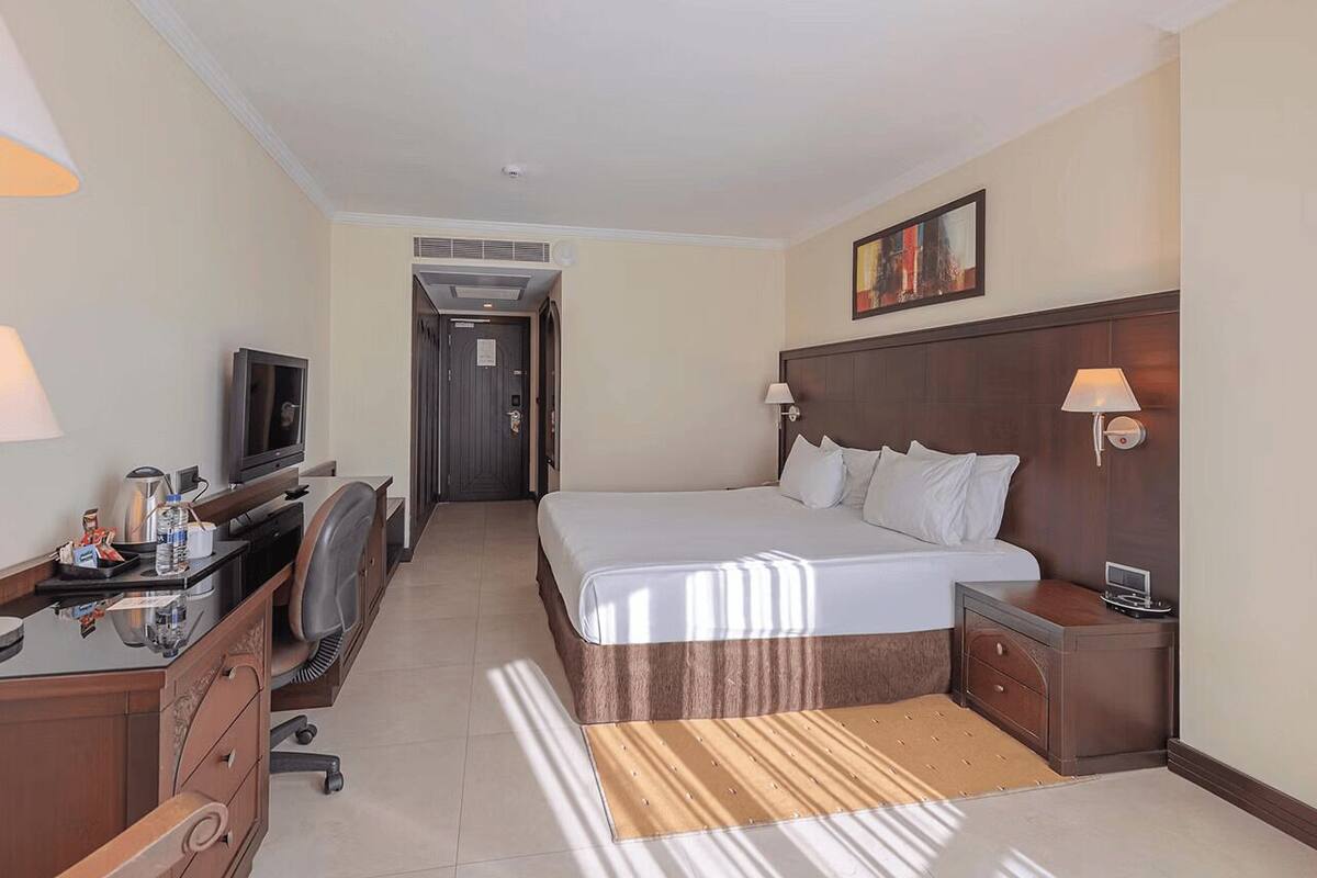 Una habitación de hotel con cama, mesitas de noche, escritorio con computadora, televisión y lámpara.