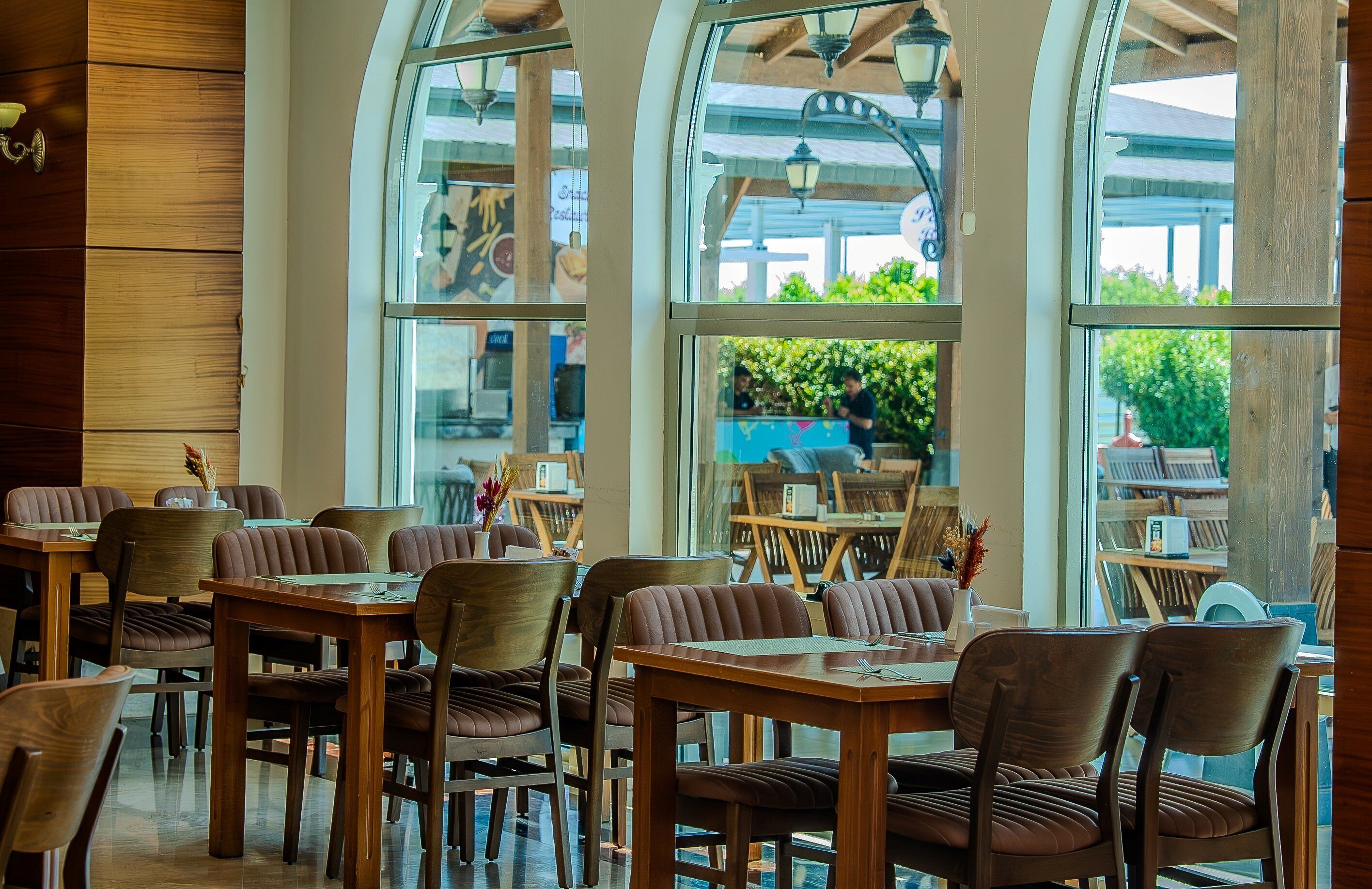 A restaurant with wooden tables and chairs, large windows, and a view of greenery outside.