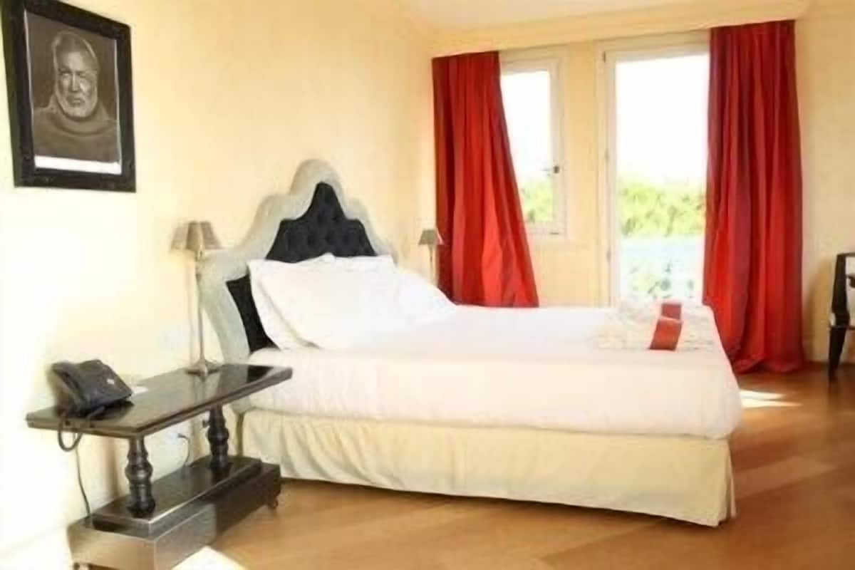 Una cama bien tendida en una habitación con piso de madera, una mesita de noche con lámpara y un cuadro enmarcado en la pared.