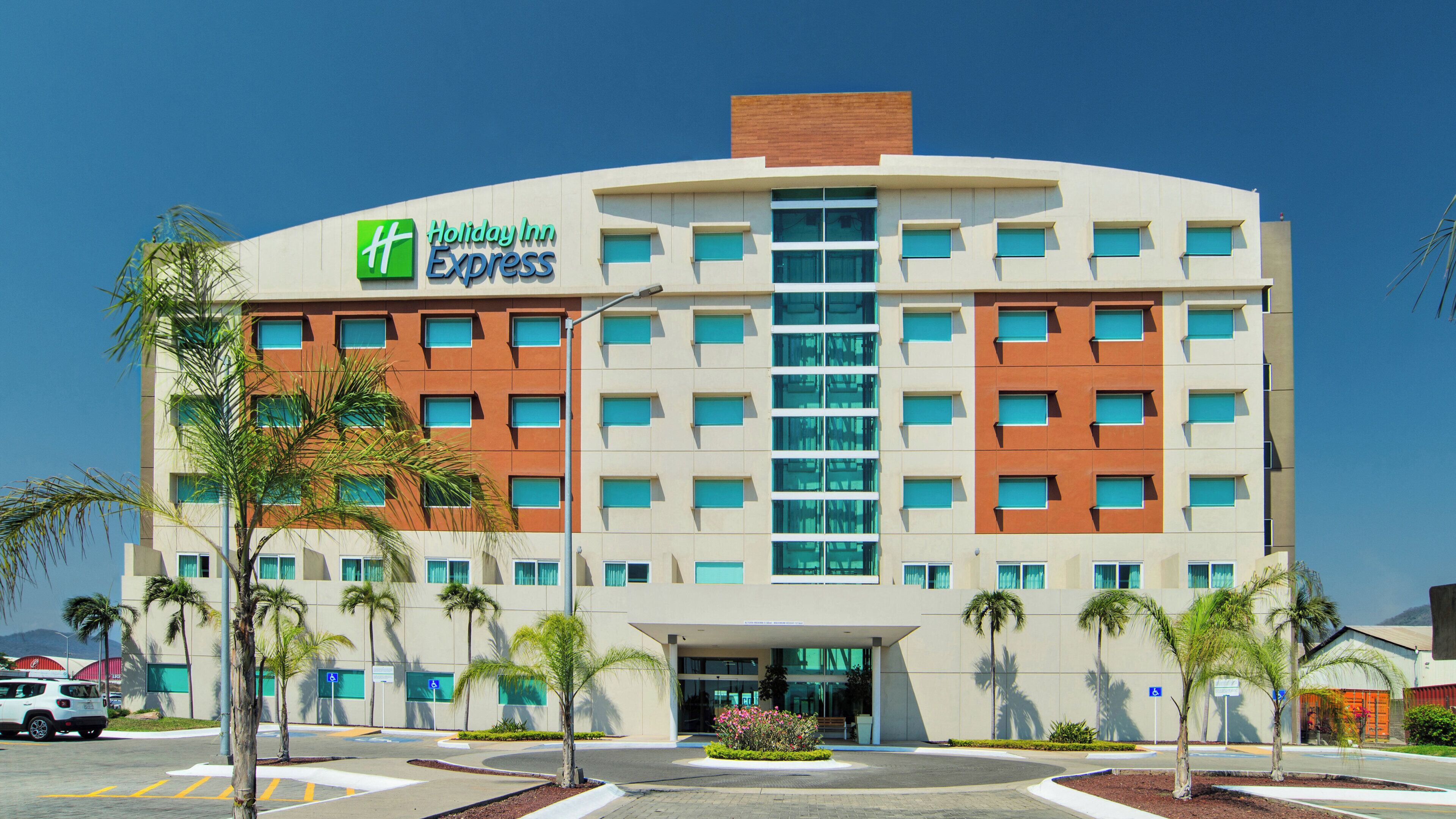A Holiday Inn Express hotel with a modern design, featuring a central entrance, multiple floors, and a parking area with palm trees.