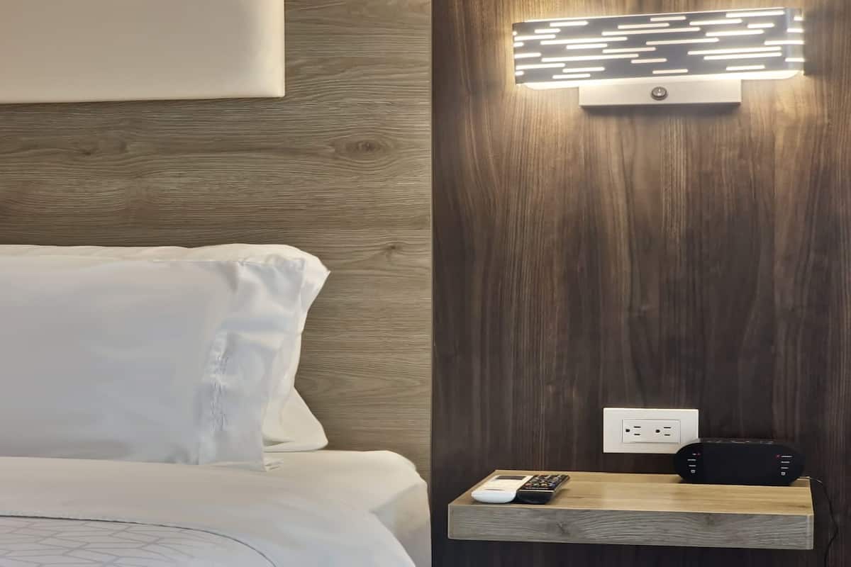 Habitación de hotel con cabecera de madera, mesita de noche con control remoto, toma de corriente e iluminación de pared.