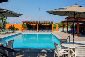 A swimming pool with lounge chairs and umbrellas, a covered seating area, and a view of the sky.