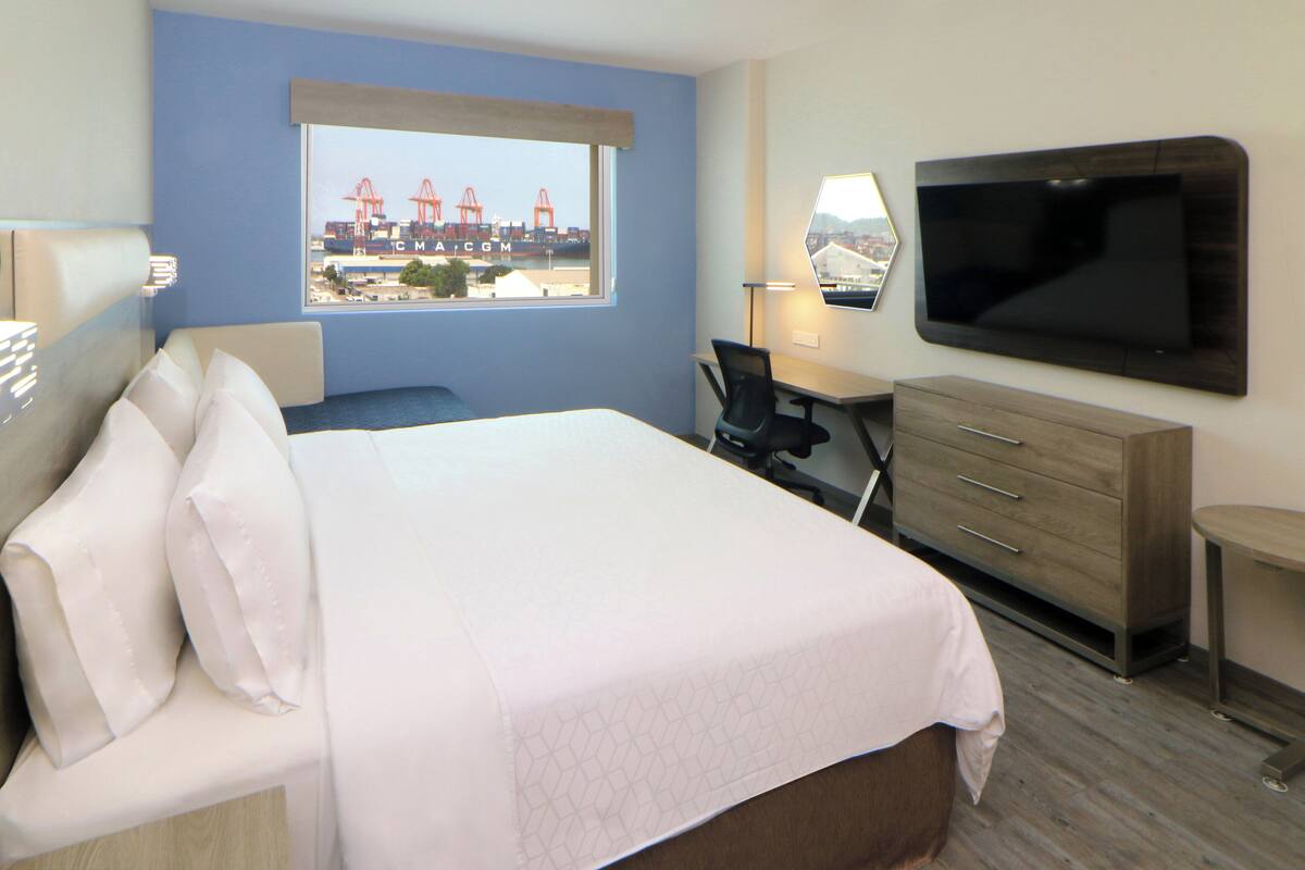 Habitación de hotel con cama, escritorio, silla, televisor y ventana con vista a grúas.