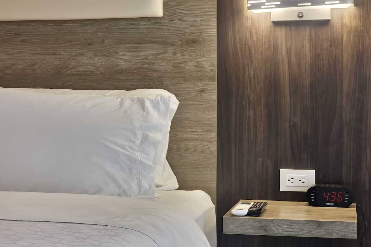 A hotel room with a bed, a nightstand with a digital clock, and a wall-mounted light fixture.