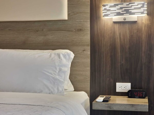 Una habitación de hotel con cama, mesita de noche con reloj digital e iluminación montada en la pared.