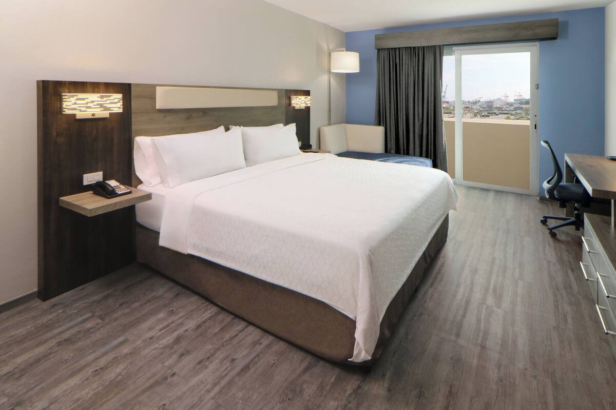 Habitación de hotel con una cama grande, un escritorio, una silla, una lámpara y vistas a la ciudad por la ventana.