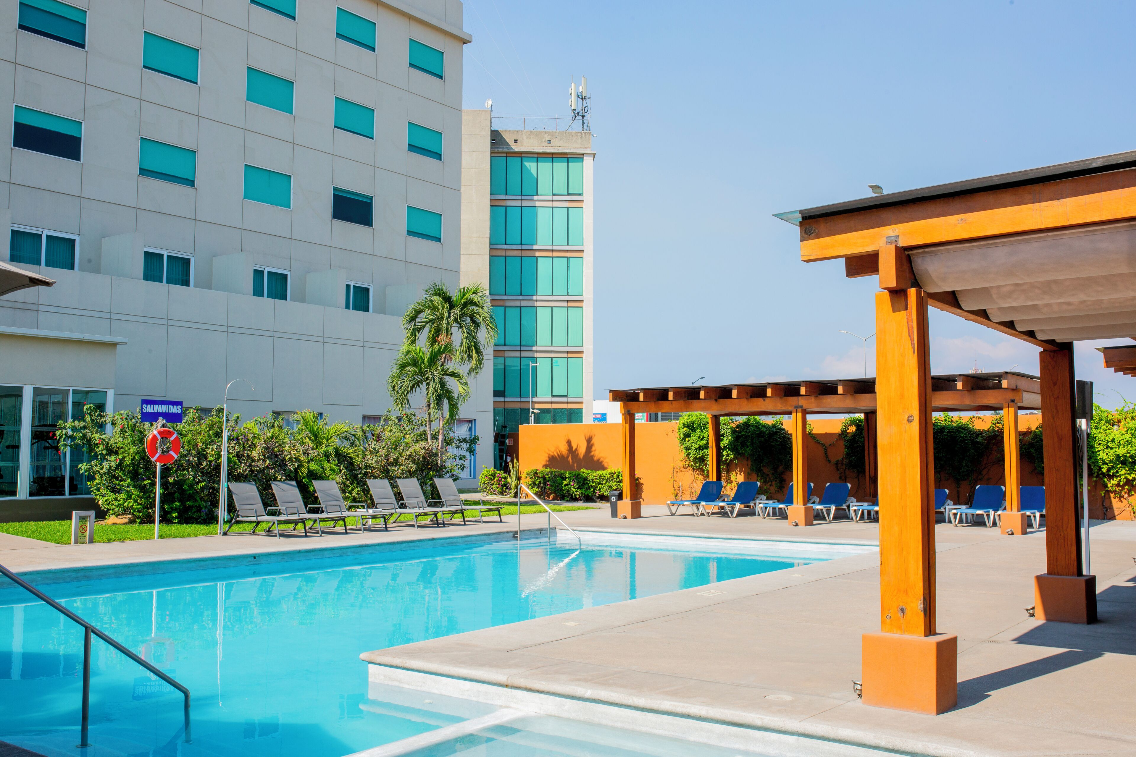 A swimming pool with lounge chairs and a wooden pergola, surrounded by a building with multiple windows and a sign reading 'SALVAVIDAS'.