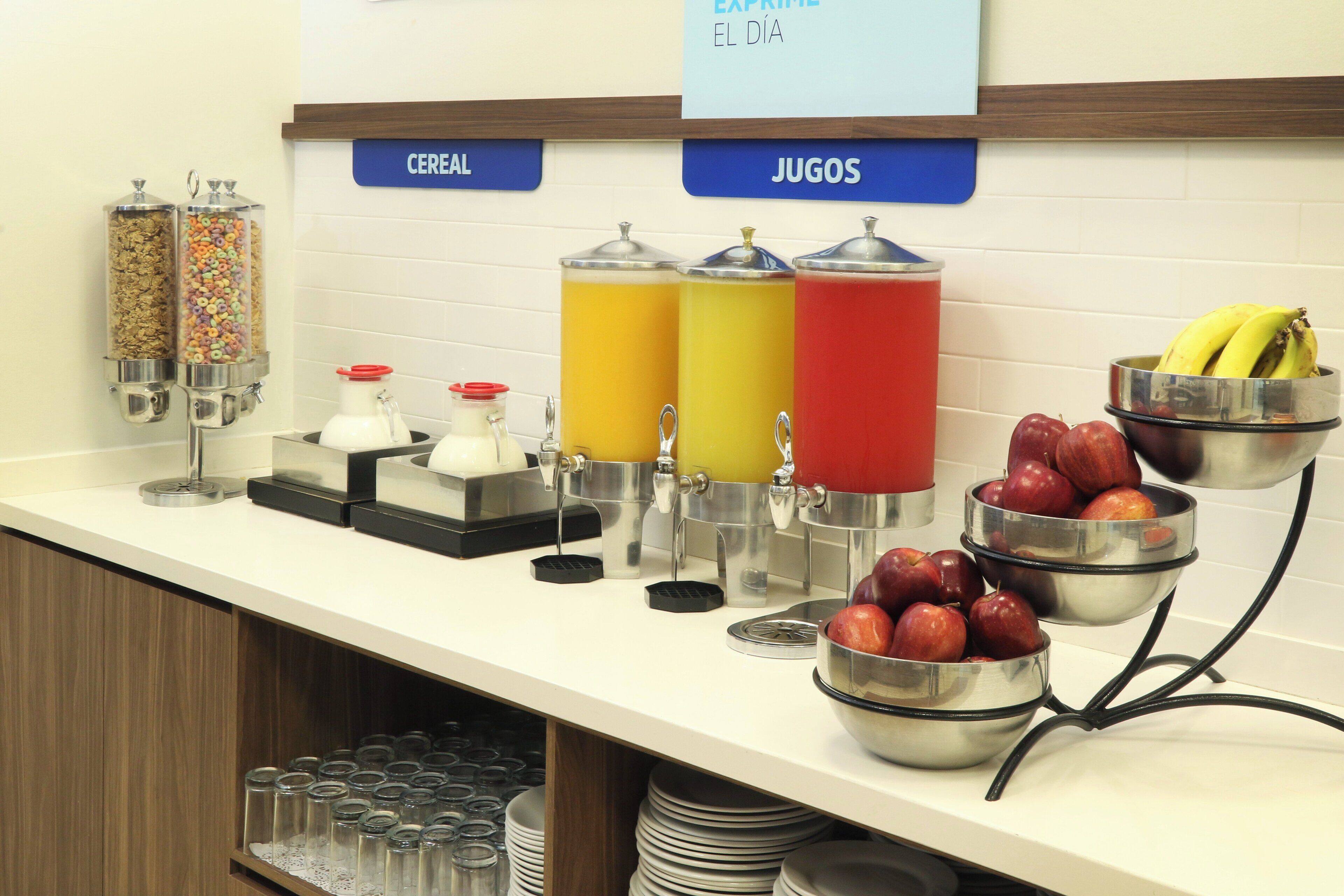 Una barra de desayuno con dispensadores de cereales y zumos, y cuencos de frutas y manzanas.
