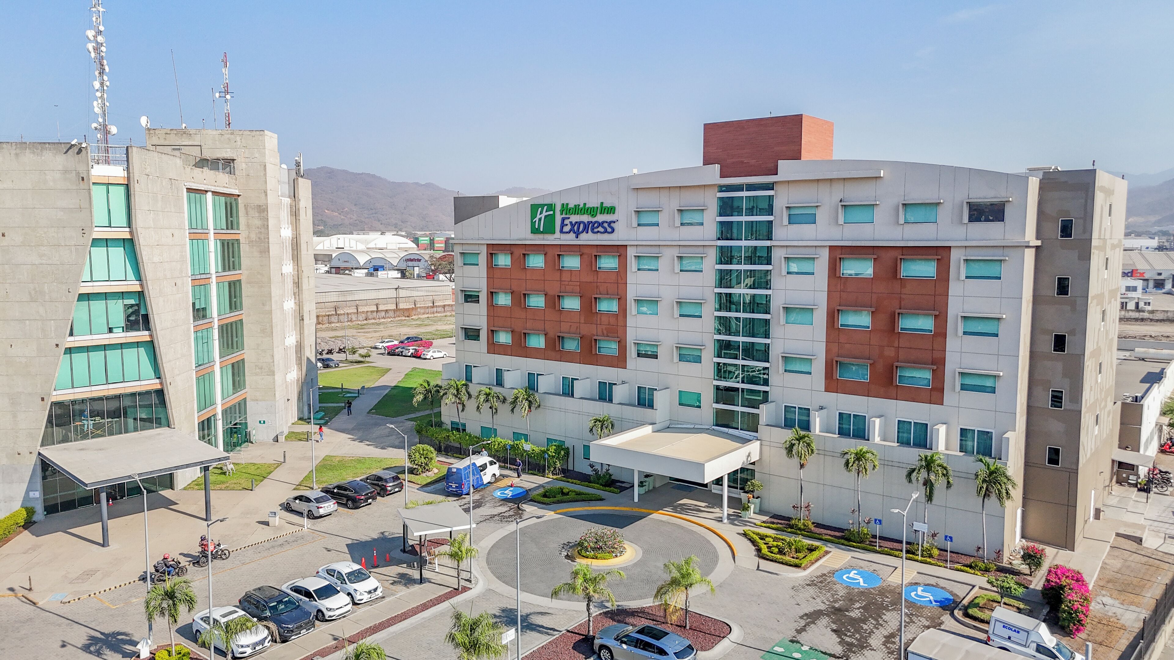A Holiday Inn Express hotel with a modern design, featuring a parking lot, a landscaped area with palm trees, and a view of the surrounding cityscape.