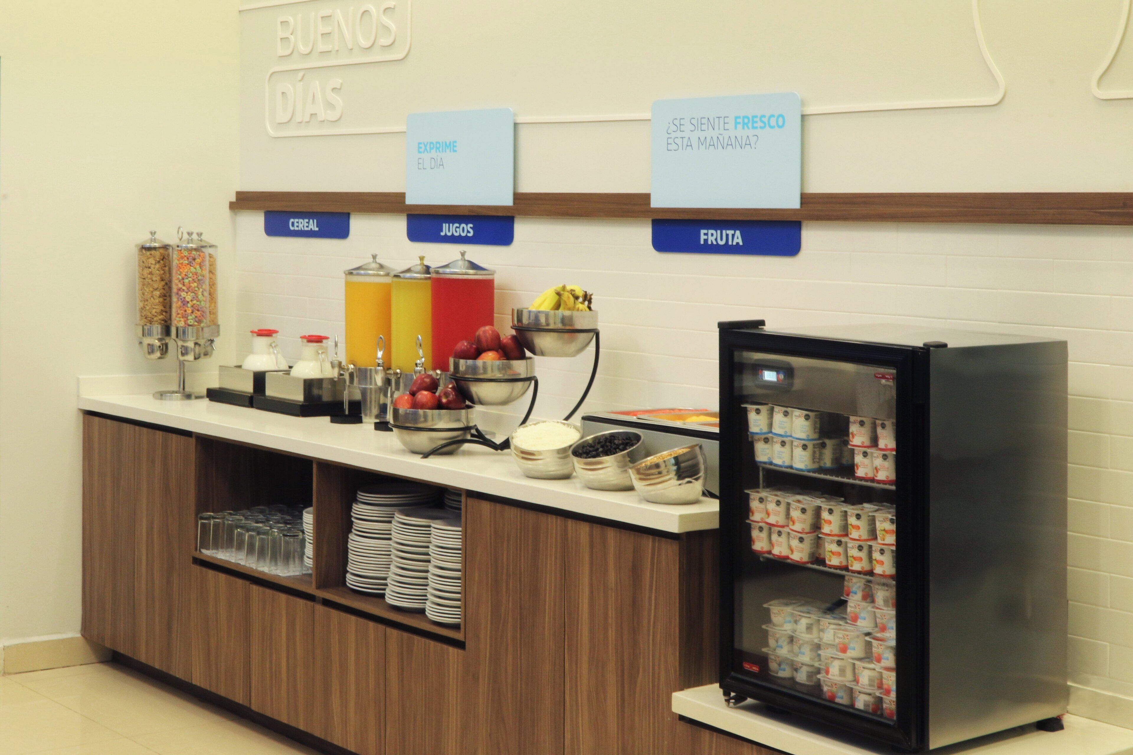 Una barra de desayuno con dispensadores etiquetados para cereales, jugos y fruta, y un refrigerador lleno de bebidas.