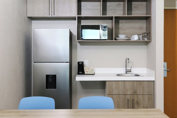 A compact kitchen with a stainless steel refrigerator, a microwave, a coffee maker, and a sink. There are two blue chairs and a wooden table. - Holiday Inn Express Manzanillo by IHG (Manzanillo)